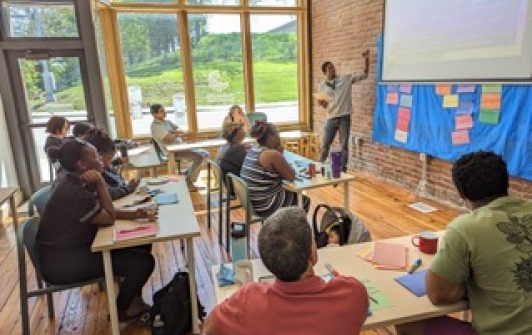 students at desks look up a teacher pointing to a projector