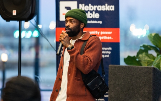A man in a green beanie and orange cardigan speaks into a microphone