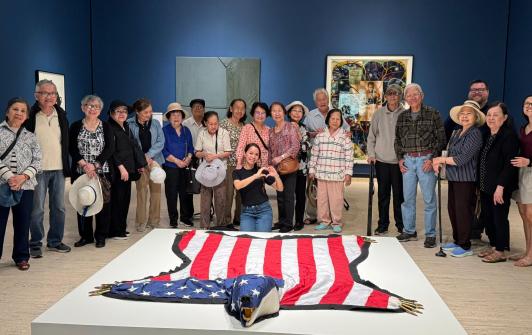 A large group of people on a field trip to the art museum pose behind a bear-shaped American flag 