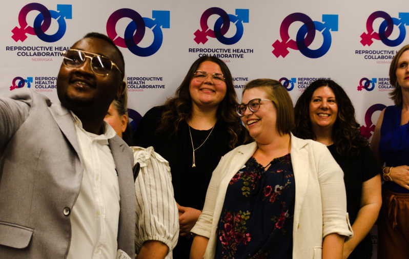 A group of four people taking a selfie, standing against a wall patterned with Reproductive Health Collaborative Nebraska's logo