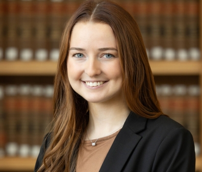 A headshot of a woman with medium-length brown hair wearing a tan shirt with a black blazer. She is standing in front of shelves of books.