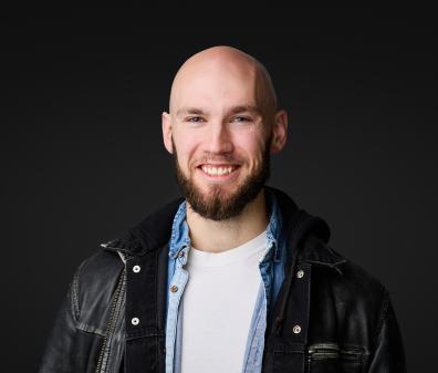 Headshot of a smiling man wearing a white shirt layered with a denim shirt and leather jacket