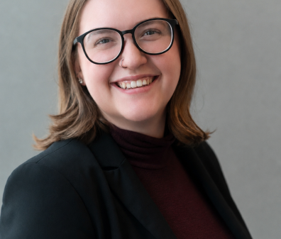 Headshot of a woman with medium length light brown hair wearing glasses, a maroon shirt, and black blazer