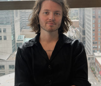 A headshot of a man with medium length hair and a black button-down shirt, against a background of city buildings