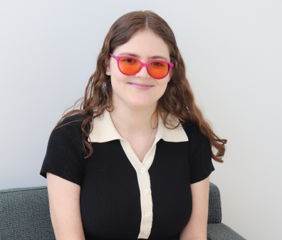 Headshot of young woman with shoulder length light brown hair wearing a black short sleeve blouse with a cream collar and pink tinted glasses