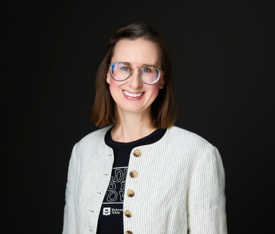 Headshot of a smiling woman with short brown hair wearing round glasses and a white striped blazed with brown buttons over a black t-shirt