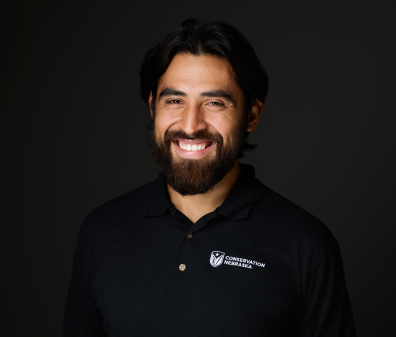 Headshot of a smiling man with dark hair and a short beard wearing a black polo stitched with the Conservation Nebraska logo
