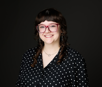 Headshot of a smiling person with brown shoulder length hair wearing red glasses and a black and white polka dot button up shirt
