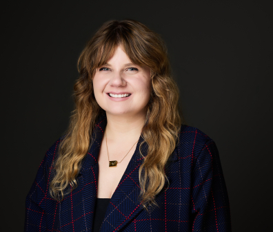 Headshot of a smiling woman with medium length blonde hair and bangs, wearing a navy and red plaid blazer